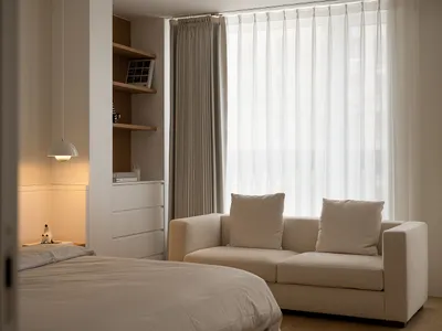 A minimalist bedroom featuring a comfortable sofa with white cushions, white curtains, and a white pendant light.