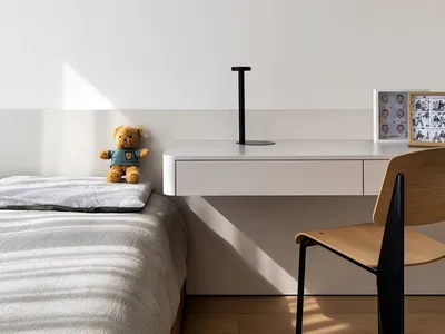 A minimalist bedroom featuring a bed with a grey blanket, a small shelf with a teddy bear and a black lamp, and a wooden chai