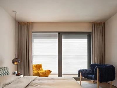 A modern bedroom featuring a beige fabric bed with a yellow and blue upholstered chair in front of a large window with blinds