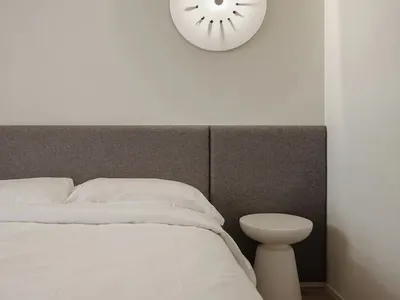 A minimalist bedroom featuring a white bed with a gray upholstered headboard against a light-colored wall, illuminated by a u