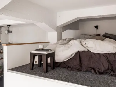 A minimalist bedroom with a bed covered in beige and brown bedding, a wooden bedside table, and concrete walls.