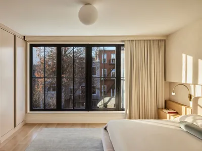 A minimalist bedroom featuring a large bed with white linens, a wooden headboard, and a large window with beige curtains and