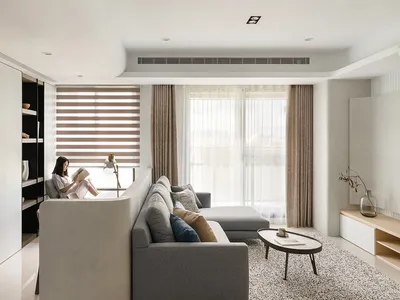 A minimalist living room with fabric curtains, concrete cabinets, and grey sofa, featuring a woman reading a book.