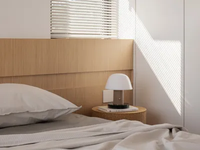 A minimalist bedroom featuring a bed with wood headboard and blinds, illuminated by sunlight casting geometric shadows.