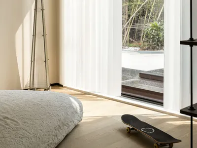 A minimalist bedroom with white curtains and a view of nature, featuring wood flooring and a skateboard.