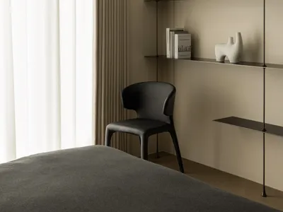 A modern minimalist bedroom with a black chair, floating shelves, and neutral tones on the walls and curtains.