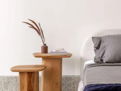 A modern minimalist bedroom with a white bed and wooden side tables and stools. The room features a vase with dried flowers o