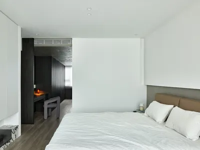 A minimalist bedroom featuring a white clothed bed, with white walls and a mix of dark and light wooden textures.