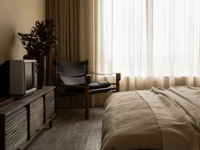A minimalist bedroom featuring a bed with neutral-toned textiles, a wooden armchair, and a vintage television on a rustic cab