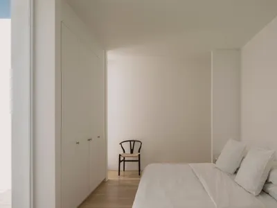 A minimalist bedroom with white walls, a wooden bed, and a simple chair.