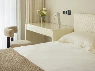 A minimalist bedroom featuring a white fabric bed against a light beige wall, illuminated by natural light filtering through