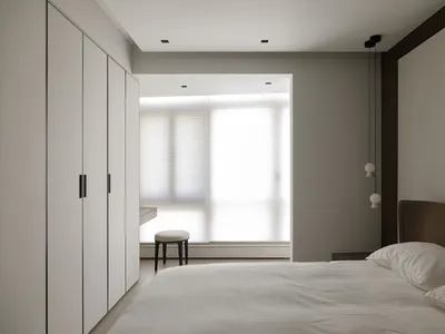 A minimalist bedroom with white and beige neutral colors, featuring a bed, a window with blinds, and a small chair in the cor