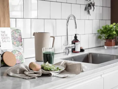 A modern kitchen with a marble countertop, featuring a glass pitcher, green smoothie, pears, and a plate with leafy greens an
