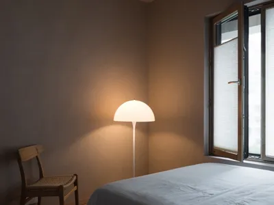 A minimalist bedroom with a wooden floor and fabric bedding, an open window with blinds, and a floor lamp illuminating the sp