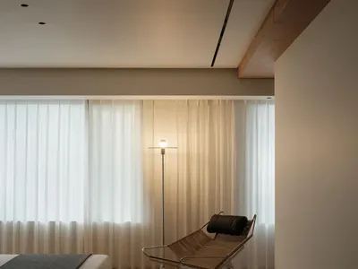 Minimalist bedroom featuring a wooden bed, glass windows, and fabric curtains, illuminated by a modern floor lamp.