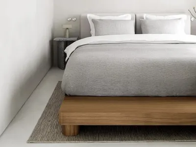 Minimalist bedroom featuring a wooden bed platform, gray and white bedding, and a textured side table with a lamp.