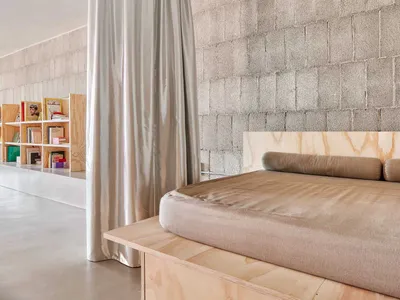 A minimalist bedroom with a fabric-covered bed on a concrete platform, featuring a concrete wall in the background and a ligh