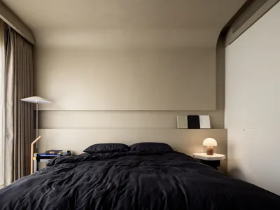 A minimalist bedroom featuring black textiles on the bed, a textured beige headboard, and neutral-toned curtains.