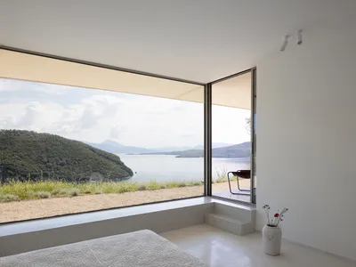 Minimalist bedroom featuring concrete flooring and walls, with large windows offering scenic views of a coastal landscape.