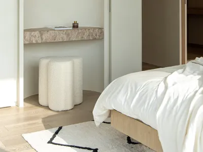 Modern minimalist bedroom with a white marble floating shelf, natural fiber stool, and white bedding.