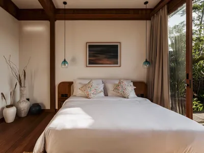 A minimalist bedroom with a wooden bed and white bedding, featuring a painting and blue pendant lights.