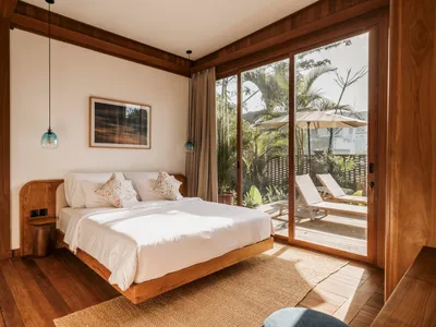 Modern bedroom interior with white floating bed on wooden floors, overlooking a patio with palm trees and lounge chairs.