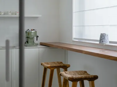 A modern minimalist kitchen with a wooden countertop and stools, featuring a green espresso machine and a window with blinds.