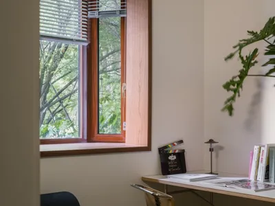 A modern home office featuring a wooden window with blinds, a desk with books and a chair, and green foliage outside the wind
