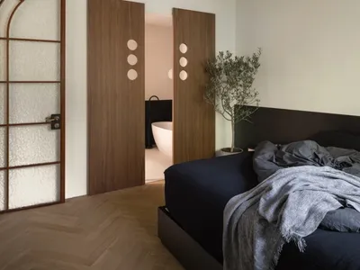A modern bedroom featuring a black bed with a dark-colored headboard and wooden floors, with a glimpse of a bathroom in the b