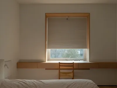 Minimalist bedroom with a floating desk and wooden chair, featuring light-colored walls and a window with a blind.