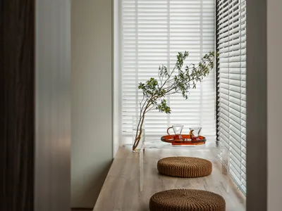 A minimalist living room with wooden furniture, clear glass vase, and white blinds.