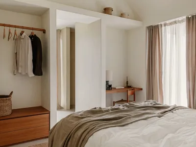A minimalist bedroom with a bed covered in linen sheets and a wooden desk under a window with sheer curtains.
