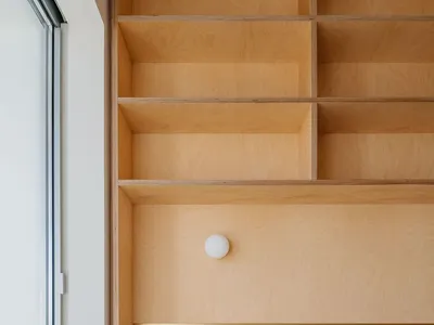 A minimalist bedroom featuring a wooden storage unit within the headboard area, next to a large window.