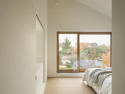 Minimalist bedroom with a large window overlooking a garden, featuring a bed with a white blanket and a wooden floor.