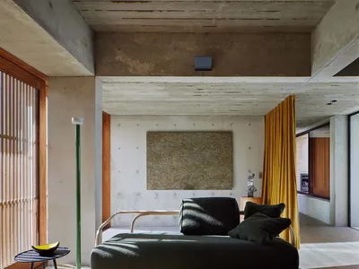 A modern industrial living room featuring bare concrete walls, a dark green lounge chair, and a yellow curtain.