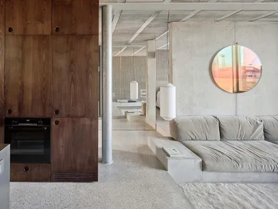 A minimalist living room with concrete walls and floor, featuring a grey sofa, a wooden cabinet, and a round window.