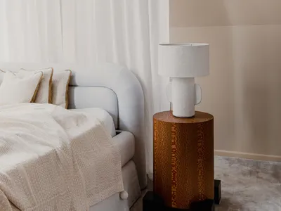 A minimalist bedroom featuring a white bed with pillows, a beige blanket, and a wooden side table with a ceramic lamp. White
