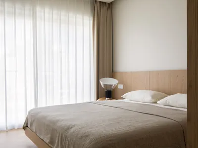 Modern minimalist bedroom with wooden furniture, white bedding, and sheer curtains.
