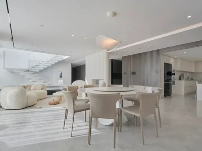 A modern living, dining, and kitchen area with white marble floors, a white round dining table surrounded by beige chairs, an