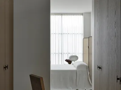 A minimalist bedroom featuring a wooden lamp, white bedding, and a curved chair.