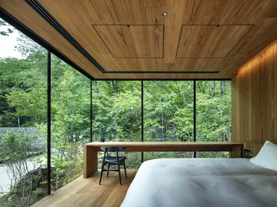 Modern minimalist bedroom featuring a white bed with a wooden table and black chair, surrounded by large glass windows overlo