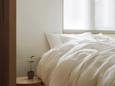 A minimalist bedroom with a bed covered in beige linen and a small wooden side table.