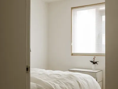 A minimalist bedroom with white walls and a wood floor, featuring a bed with a white comforter and a small bedside table with