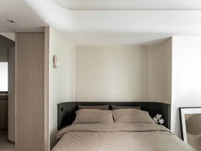 A minimalist bedroom featuring a bed with a black upholstered headboard and beige bedding, alongside a wooden headboard panel