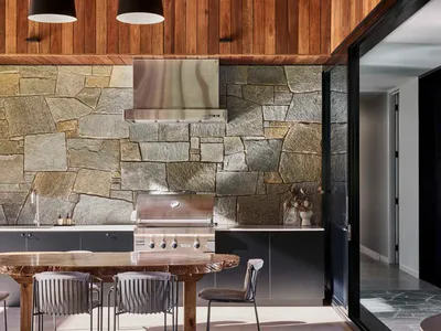 A modern kitchen with stone and concrete walls, a teak wood table, and a stainless steel grill and hood.