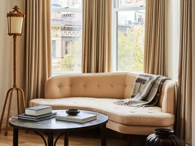 A cozy living room with a curved fabric couch, floor lamp, coffee table, and large windows draped with brown curtains.