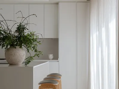 A minimalistic white kitchen with clean lines, modern stools, and large windows.