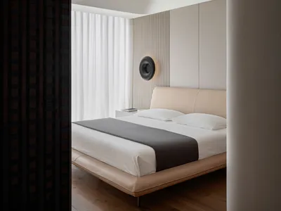 A minimalist bedroom with a beige upholstered bed, wooden flooring, and a round sconce light on the wall.