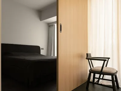 A modern minimalist bedroom with a dark bed, white curtains, and a wooden sliding door.