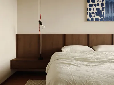 A minimalist bedroom featuring a sleek, floating dark wood nightstand with a hanging light fixture above and a large abstract